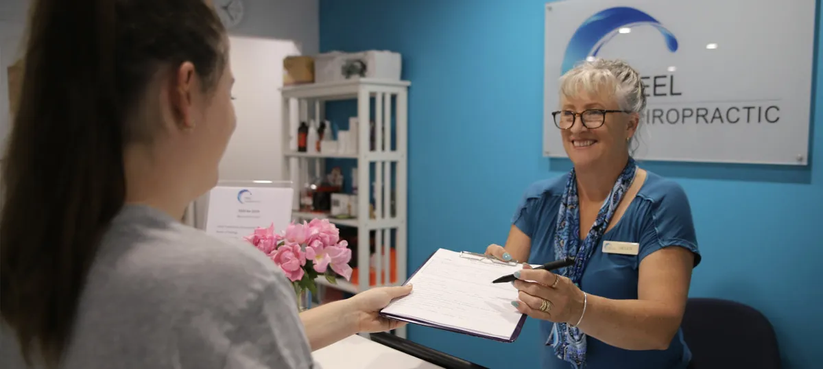 A Peel Chiropractic assistant hands a new patient the paperwork on a clipboard at reception